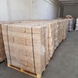 Skladowane na paletach, owinięte folią pakiety brykietu drzewnego w magazynie, widoczne worki z pelletem w tle.