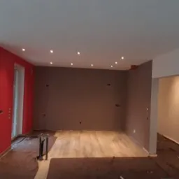 Pokój w trakcie prac wykończeniowych: świeżo pomalowana, intensywnie czerwona ściana kontrastuje z szarą ścianą, widoczne punkty świetlne w suficie, zabezpieczona podłoga i drabina malarska w rogu...