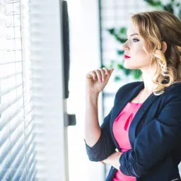 Profesjonalnie ubrana kobieta w czarnej marynarce i różowej sukience, z elegancko upiętymi blond włosami, spogląda przez okno z żaluzjami, w tle widoczna zielona roślina.