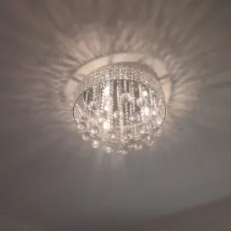 Kryształowy żyrandol sufitowy z okrągłą, chromowaną podstawą i licznymi szklanymi kulkami, rzucający dekoracyjne cienie na jasny sufit.