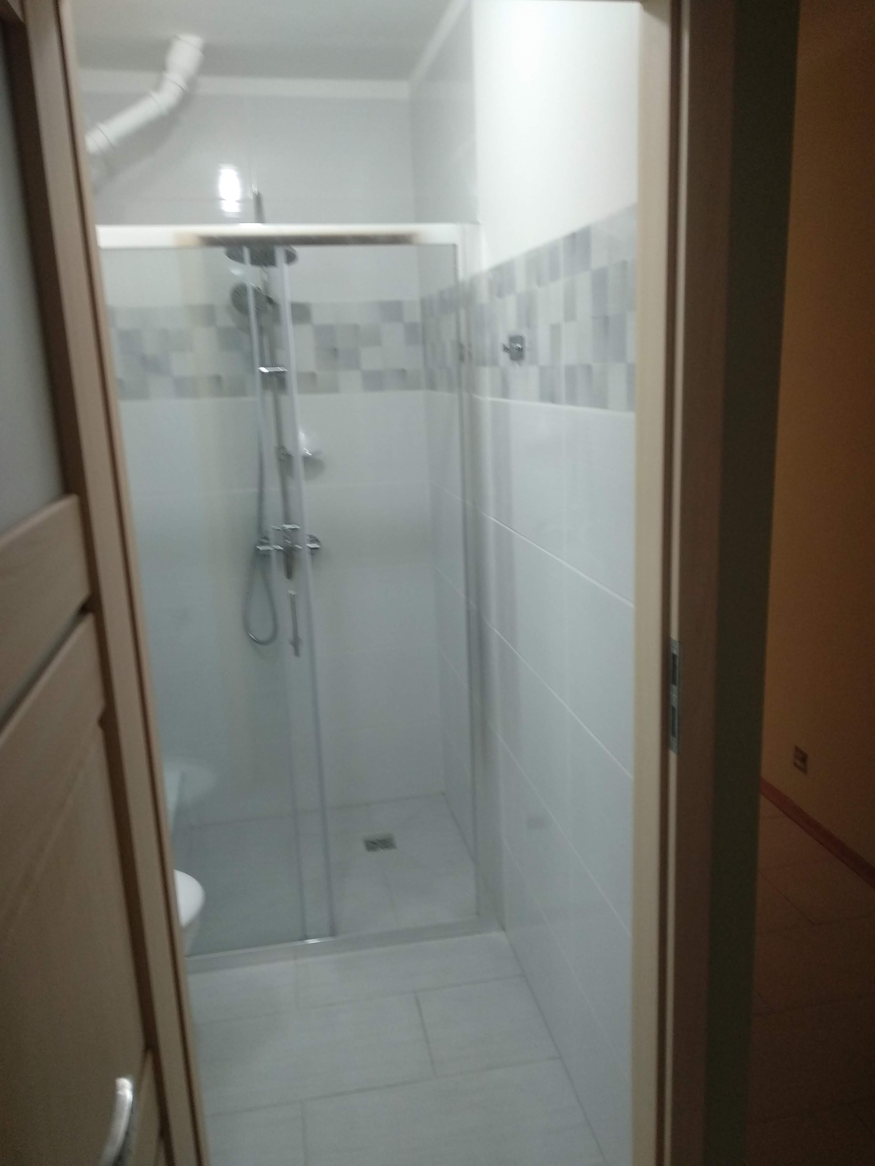 Nowoczesna kabina prysznicowa z szklanymi drzwiami, wykończona białymi i szarymi płytkami, widok zza uchylonych drzwi łazienki.