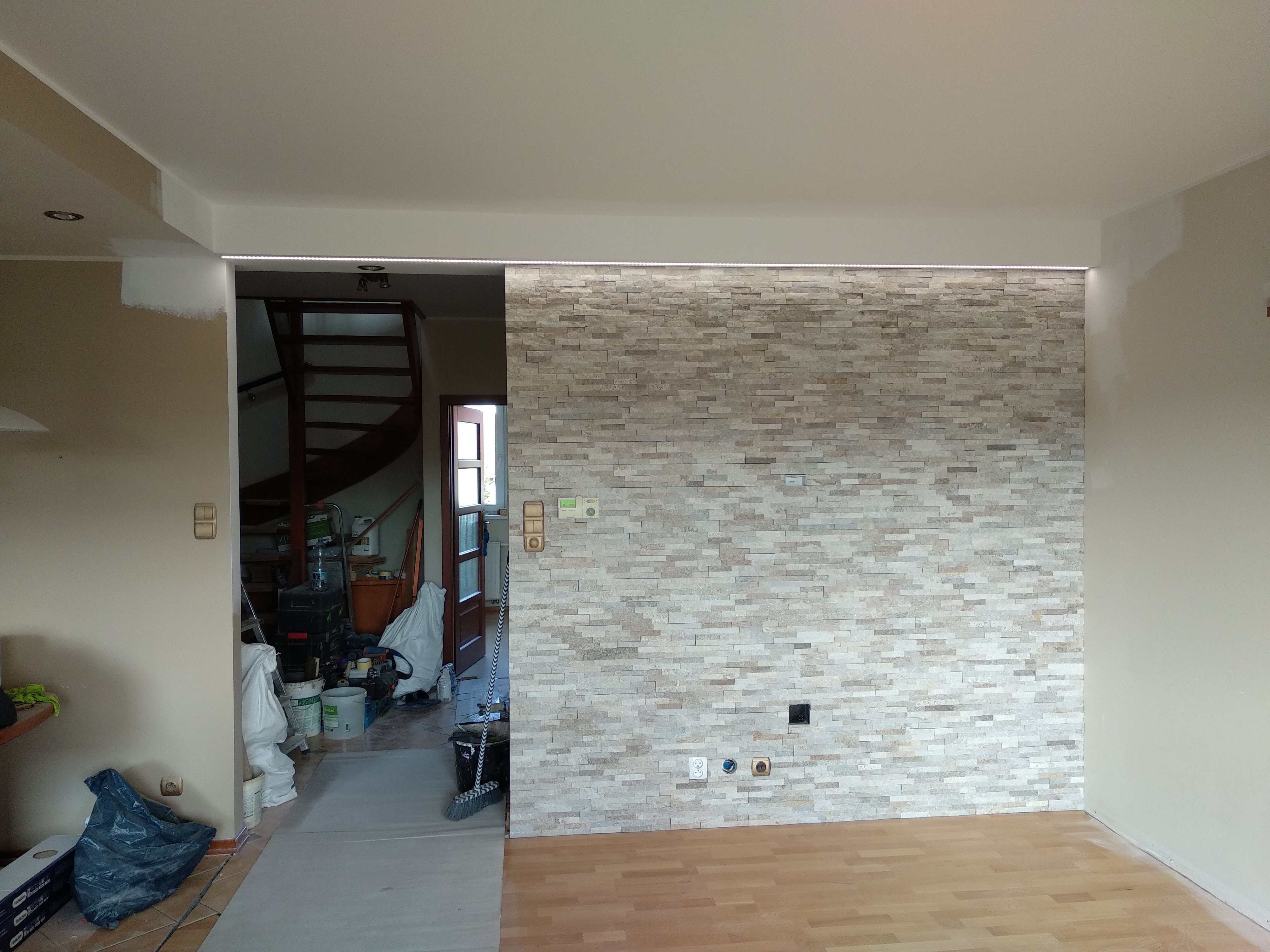 Ściana wykończona płytkami imitującymi kamień, z widocznymi gniazdkami elektrycznymi i oświetleniem LED podsufitowym, fragment schodów w tle, pomieszczenie w trakcie prac wykończeniowych.