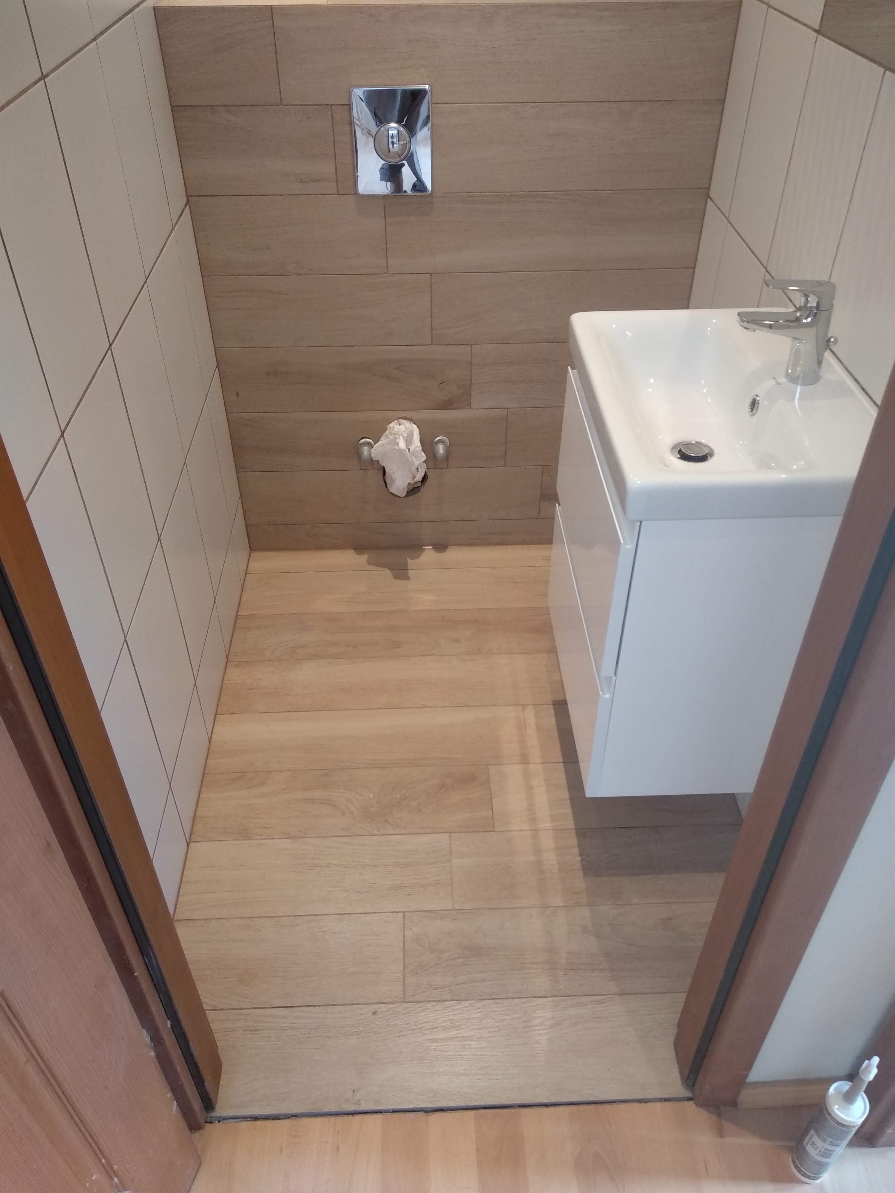 Mała łazienka po remoncie: beżowe płytki imitujące drewno na podłodze i ścianie, biała umywalka z szafką, chromowana bateria i przycisk spłuczki, widok zza framugi drzwi.