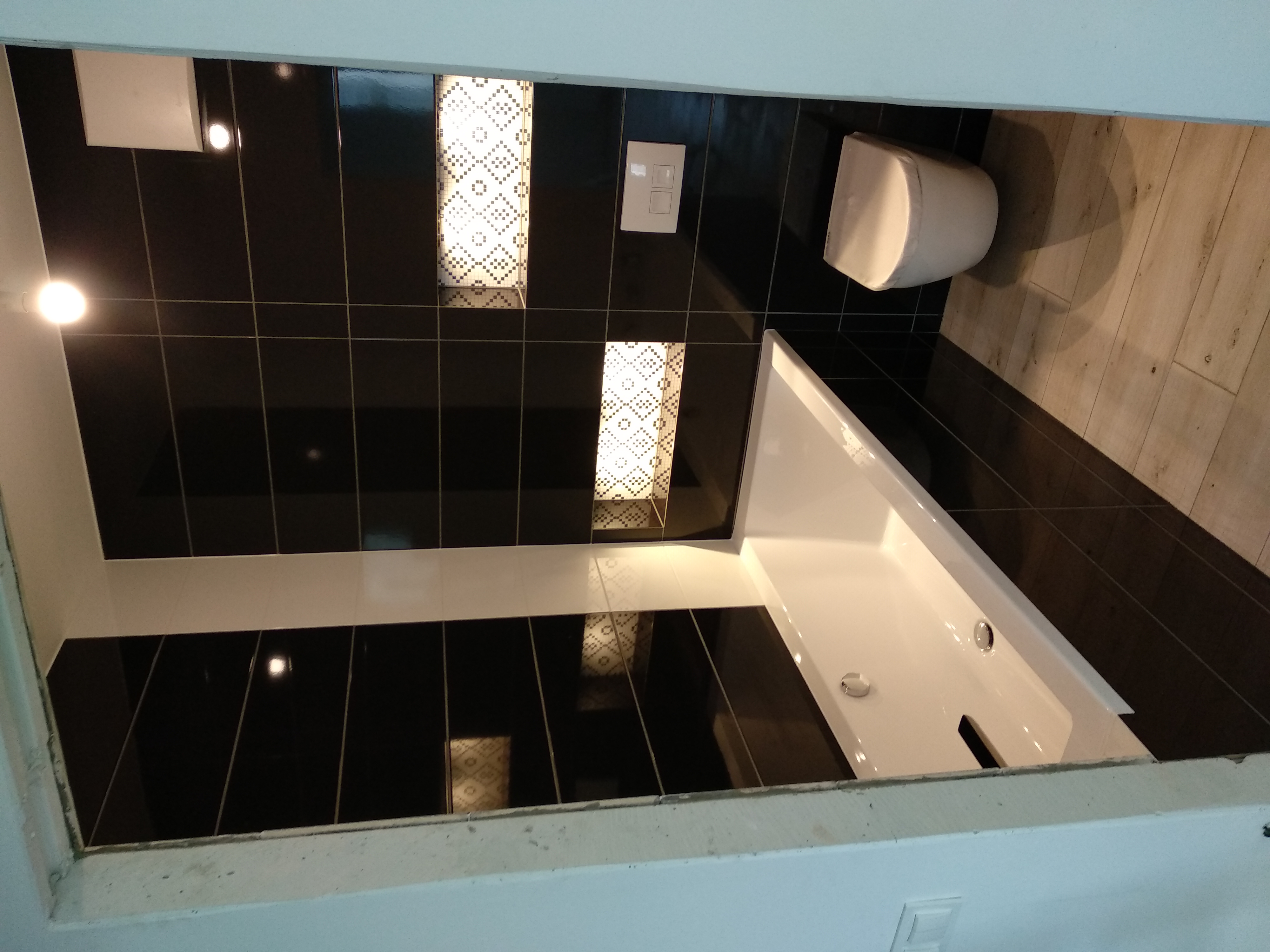 Nowoczesna łazienka z czarnymi płytkami, dekoracyjnymi wstawkami mozaiki, białą wanną i podwieszaną toaletą, wykończona drewnopodobnymi panelami podłogowymi.