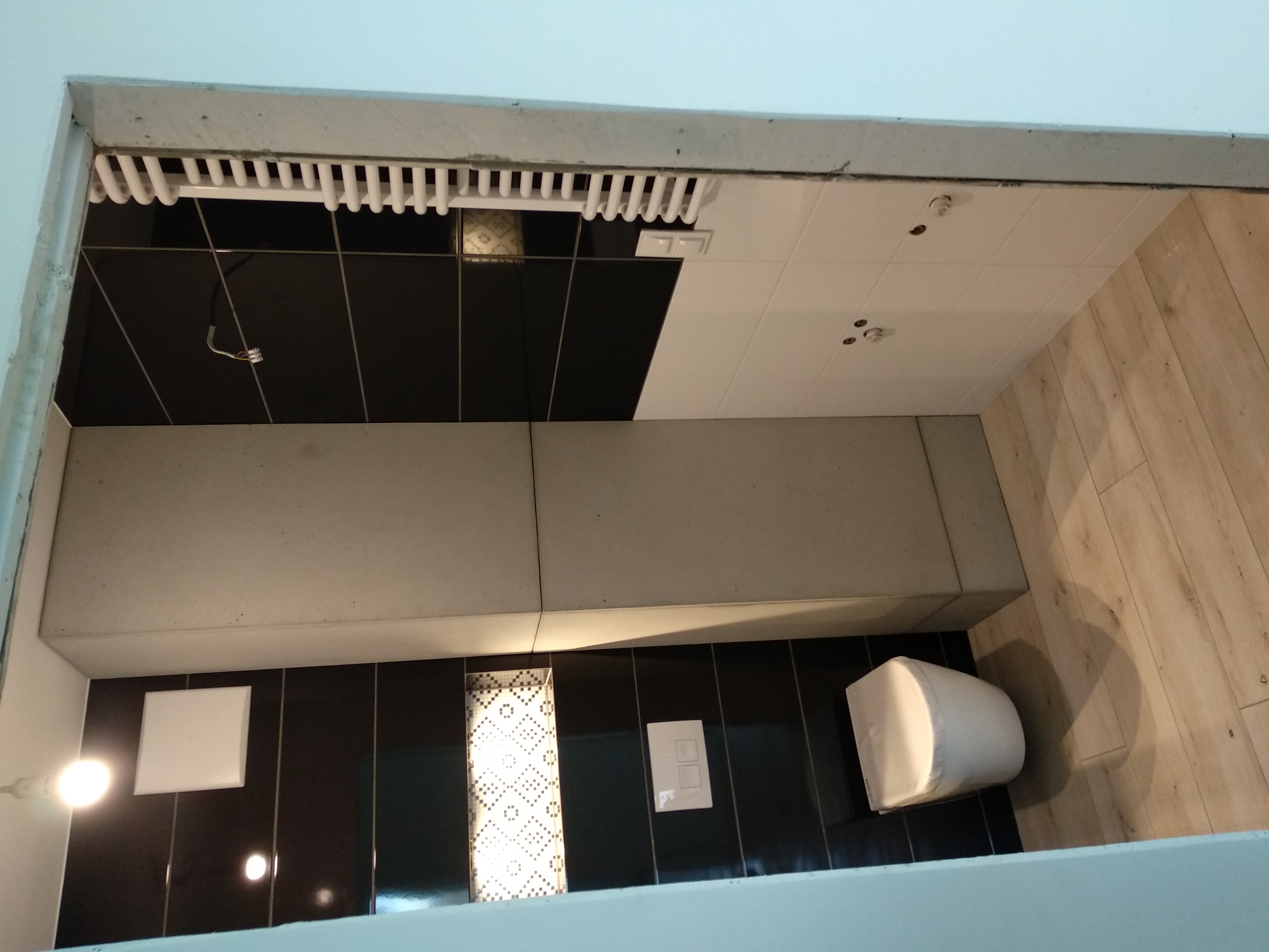 Nowoczesna łazienka z czarno-białymi płytkami, podwieszaną toaletą i drewnopodobną podłogą, widoczny fragment sufitu z elementami instalacji.