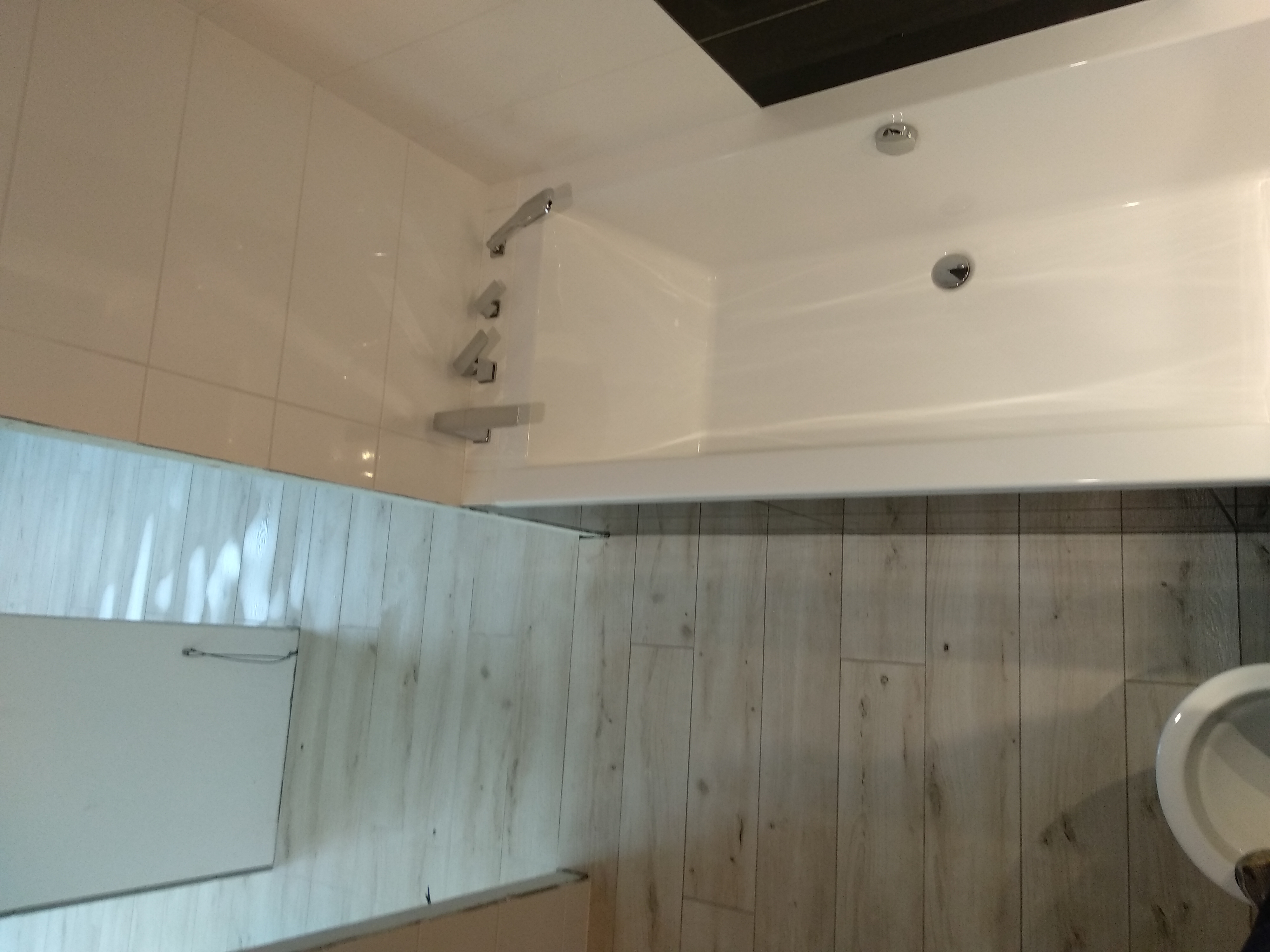 Nowoczesna łazienka po remoncie: biała wanna z chromowaną armaturą, białe płytki ścienne i podłoga imitująca drewno.