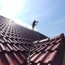 Mycie dachu z czerwonej dachówki ceramicznej pod wysokim ciśnieniem, widoczny pracownik z lancą natryskową i wężem, w tle błękitne niebo i zieleń drzew
