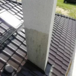 Częściowo oczyszczony komin z szarego tynku na brązowym dachu z dachówki, widoczna różnica w kolorze między czystą i brudną powierzchnią, w tle zieleń i czarny samochód.