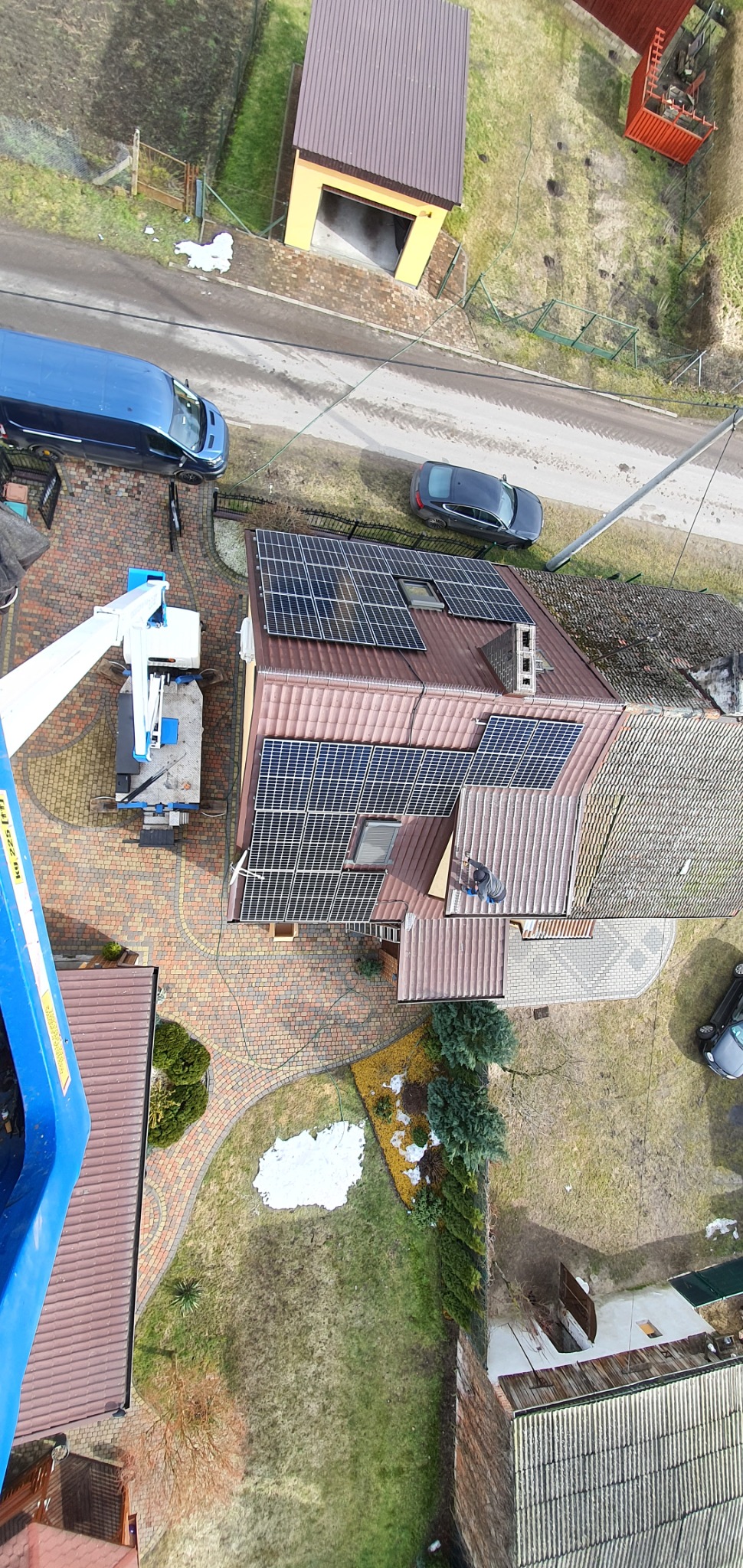 Widok z góry na dom z panelami słonecznymi na dachu, z pracownikiem na dachu i podnośnikiem koszowym obok, na tle ogrodu i budynków gospodarczych.