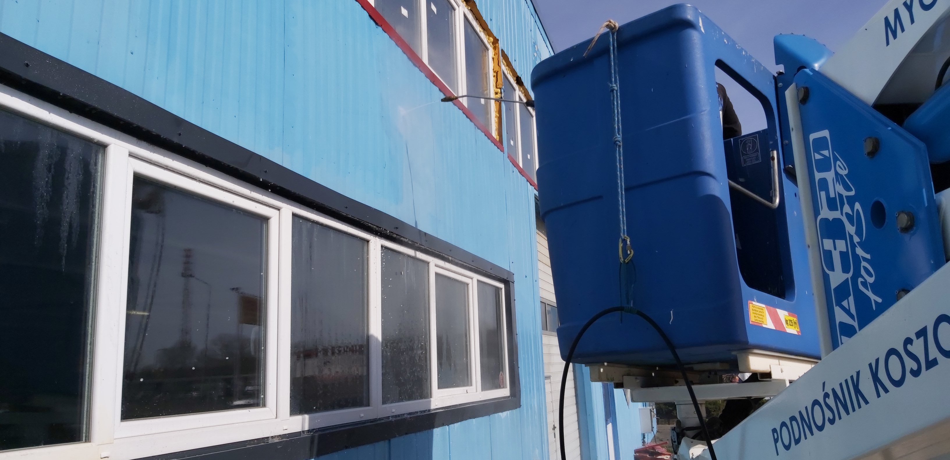 Niebieska elewacja budynku przemysłowego czyszczona myjką ciśnieniową z podnośnika koszowego; widoczne okna z czarnymi obramowaniami i ślady wody spływającej po szybach.
