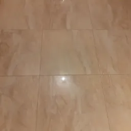 Beżowe płytki podłogowe z marmurkowym wzorem ułożone w rzędach, widoczny fragment brązowych drzwi po lewej stronie.