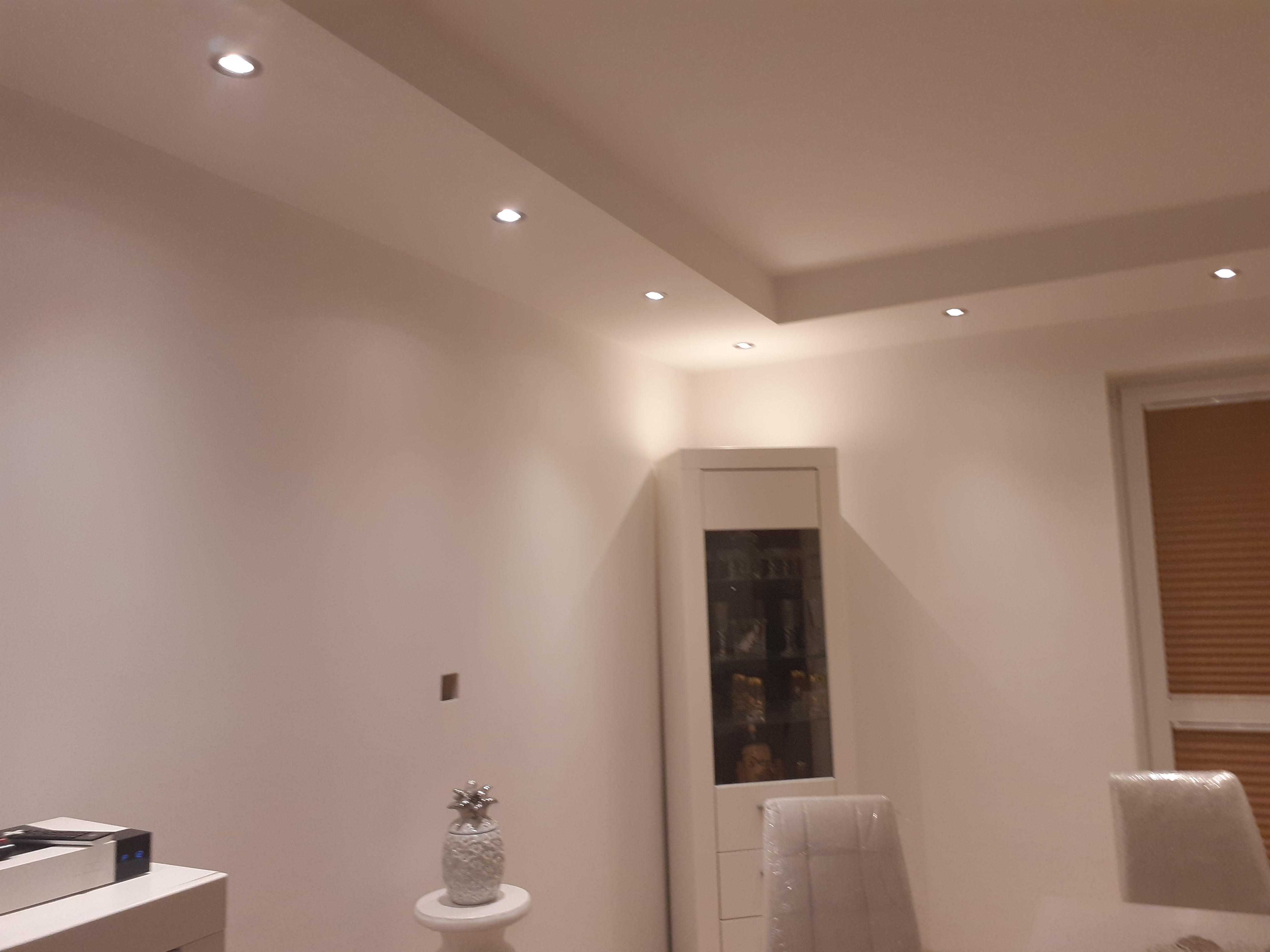 Jasne wnętrze po remoncie z punktowymi lampami w suficie, białe ściany, przeszklona witryna z dekoracjami, krzesła zabezpieczone folią, dekoracyjna figurka ananasa.