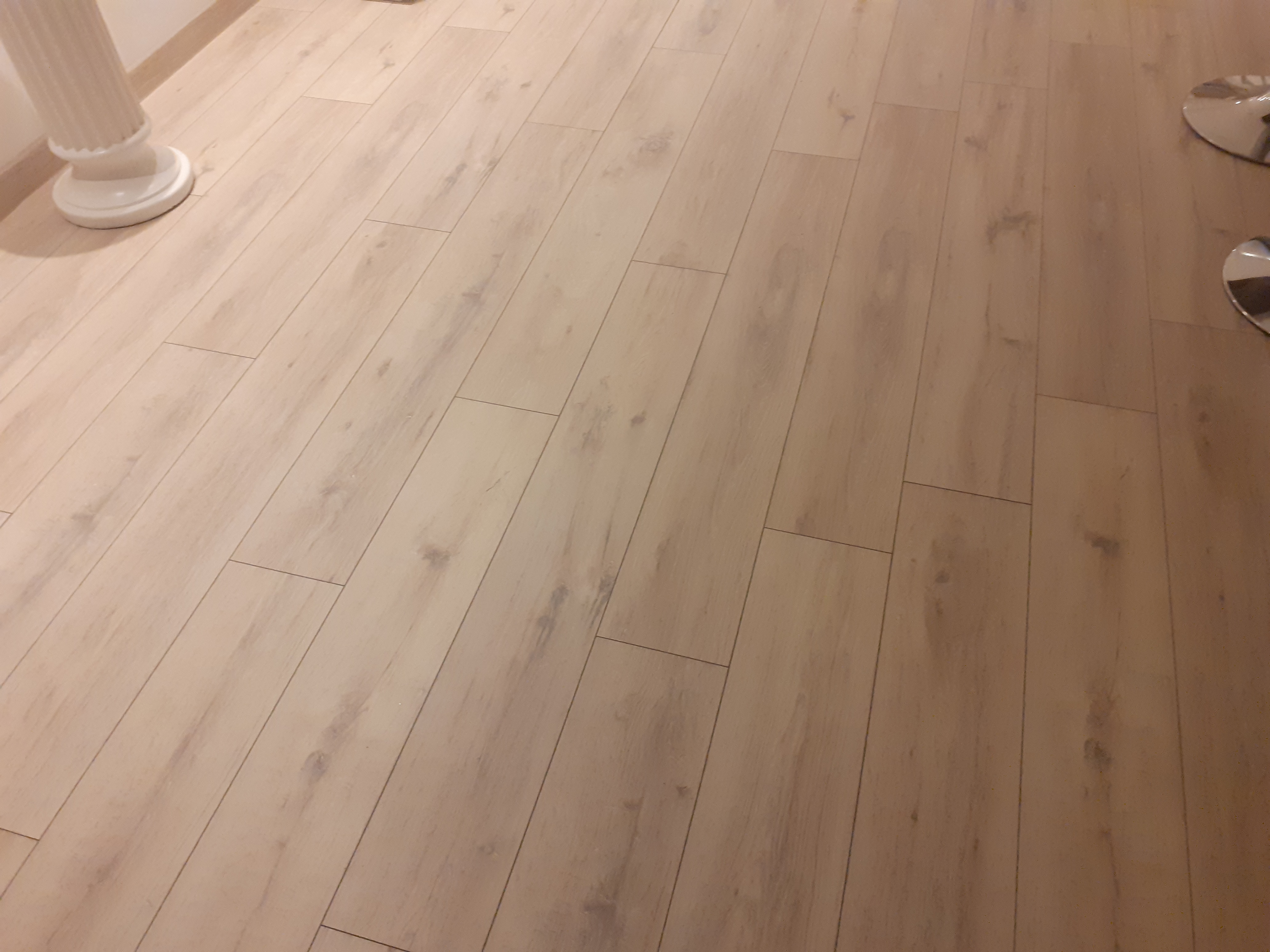 Ułożona podłoga z paneli imitujących jasne drewno, widoczna kolumna dekoracyjna i fragment metalowej nogi mebla.