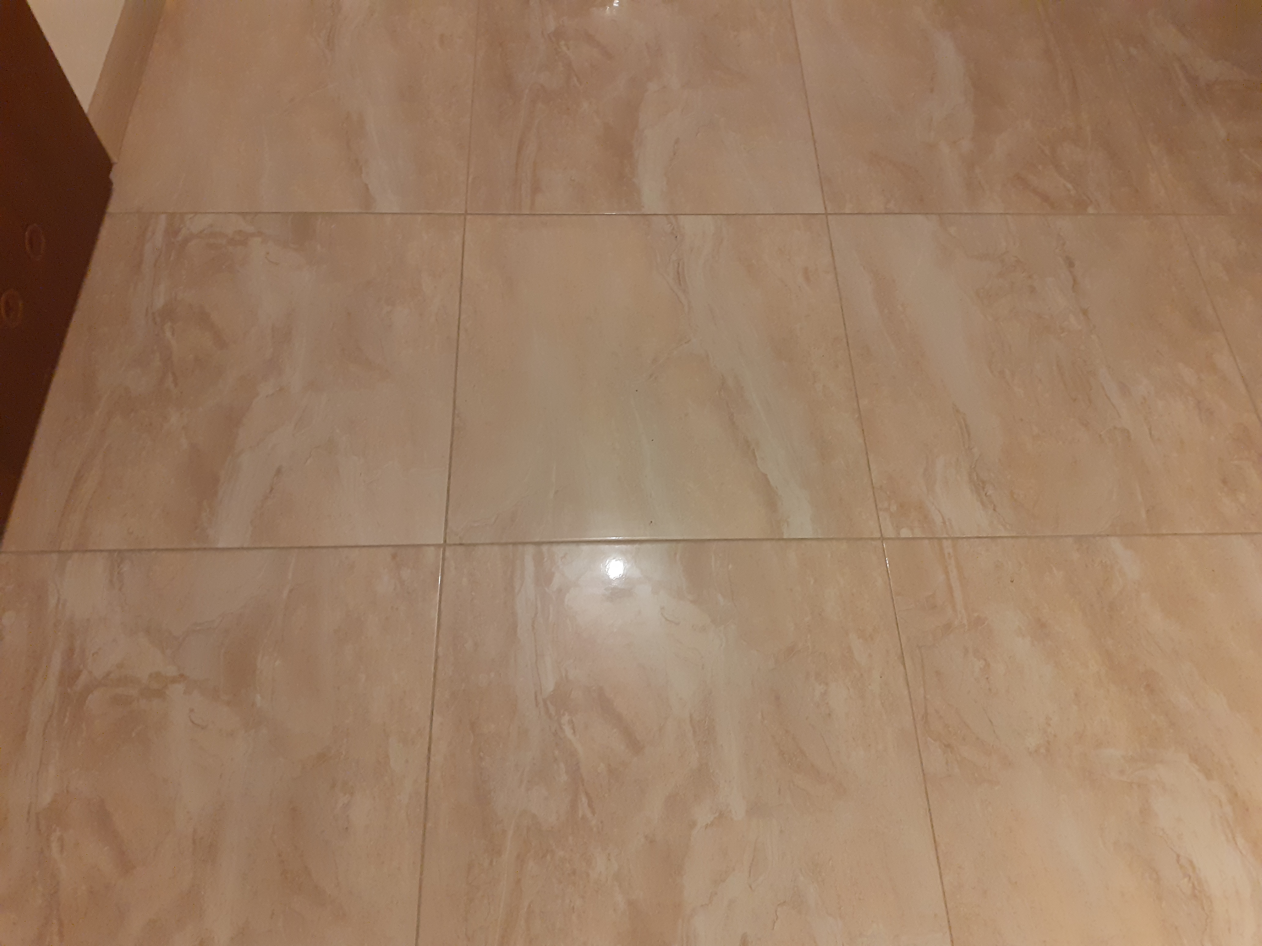 Beżowe płytki podłogowe z marmurkowym wzorem ułożone w rzędach, widoczny fragment brązowych drzwi po lewej stronie.