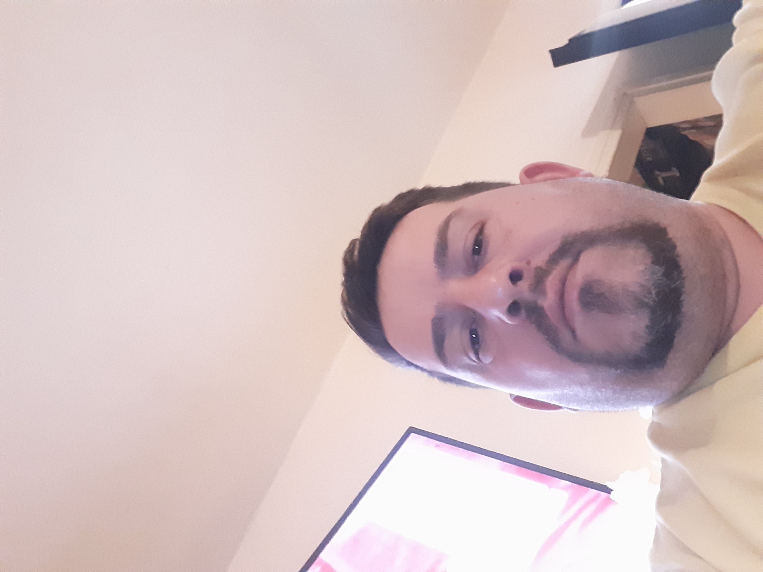 Portret mężczyzny z ciemnymi włosami i brodą, sfotografowanego z dołu, z fragmentem białego sufitu i telewizora w tle. Ujęcie z osobistej perspektywy.