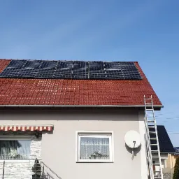 Panele słoneczne na czerwonym dachu domu jednorodzinnego z aluminiową drabiną opartą o ścianę i anteną satelitarną