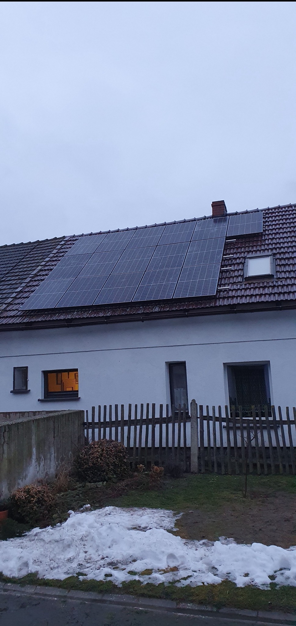 Biały dom z brązowym dachem i panelami słonecznymi, widok z dołu w pochmurny dzień, resztki śniegu na trawniku.