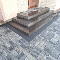 Schody wejściowe wyłożone ciemnymi, lśniącymi kostkami, otoczone szarą kostką brukową o nieregularnym wzorze, z workiem na gruz w tle.