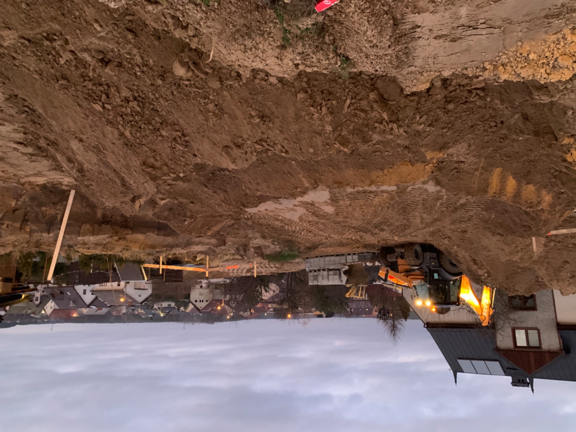 Wykop fundamentowy z widocznymi deskami szalunkowymi i koparką CASE z włączonymi światłami na tle zabudowy jednorodzinnej o zmierzchu.