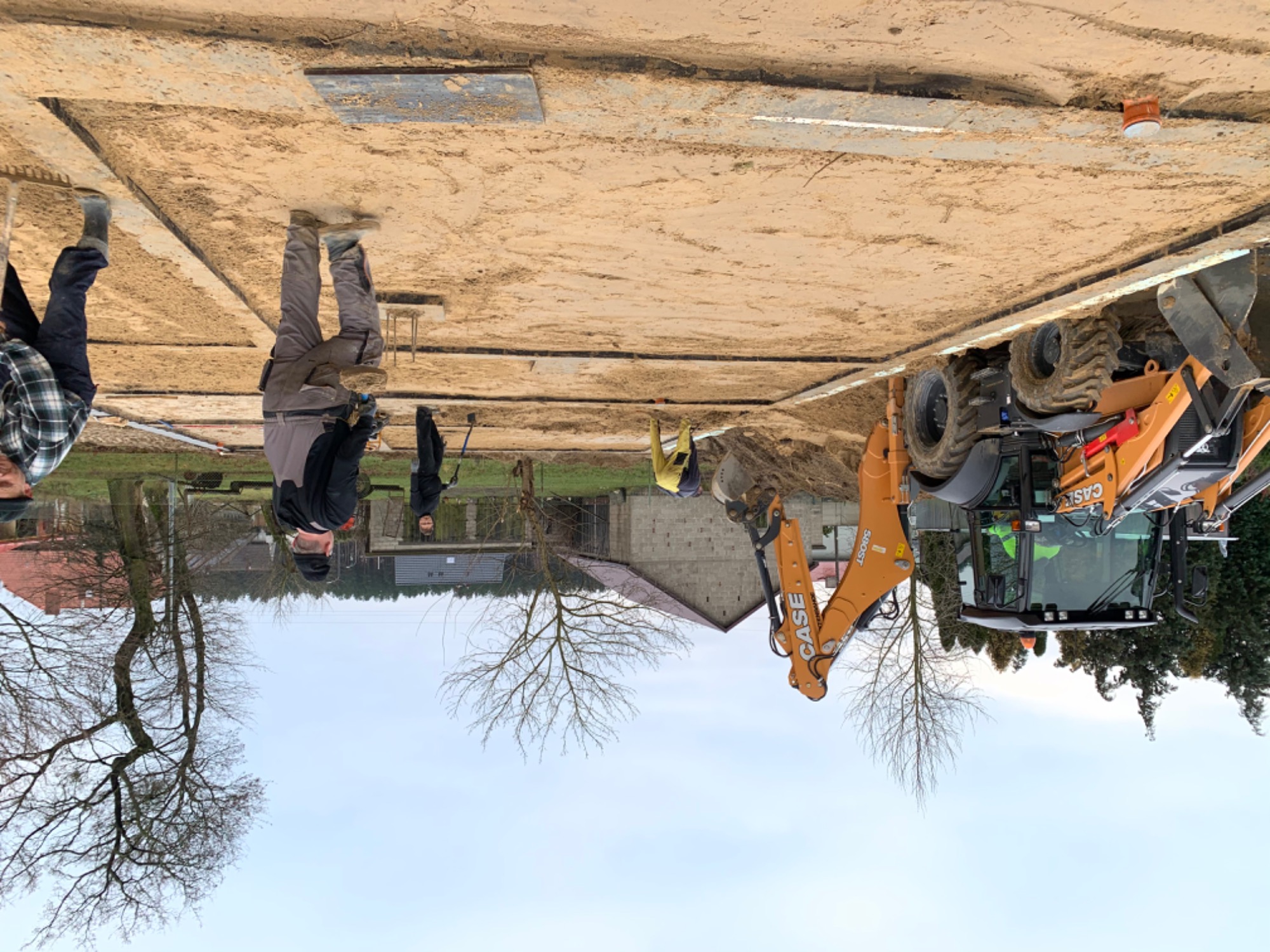 Przygotowanie terenu pod fundamenty: pracownicy z łopatami i koparka Case 580ST na placu budowy, widoczne wyznaczone granice fundamentów i podsypka piaskowa.