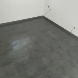 Wykończona podłoga w łazience z ciemnoszarych, matowych płytek; widoczne wystające kable elektryczne ze ściany i okno w tle.