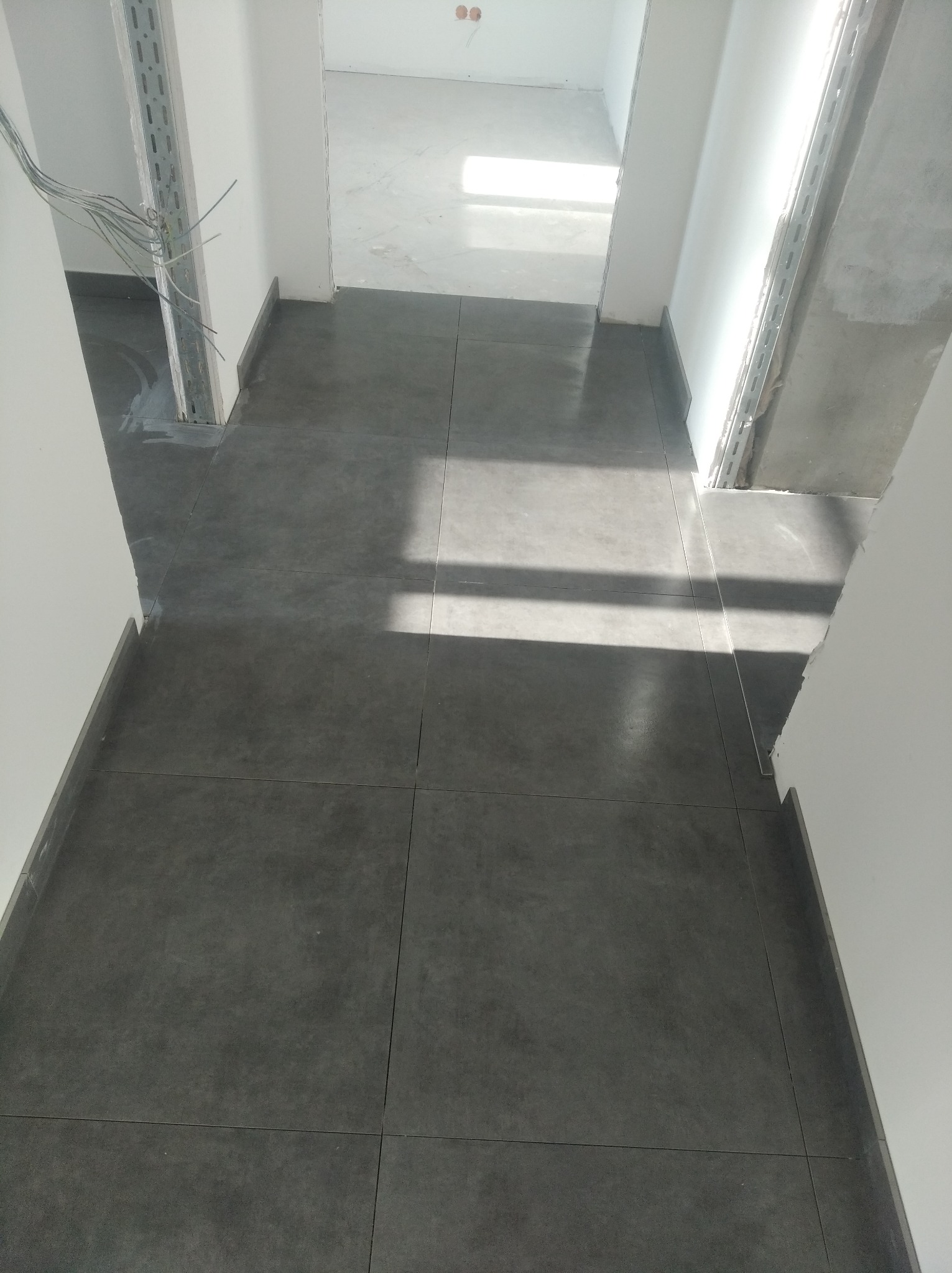 Ułożona, ciemnoszara podłoga z dużych kwadratowych płytek w nowo wykończonym korytarzu z białymi ścianami i surowym betonem na filarach.