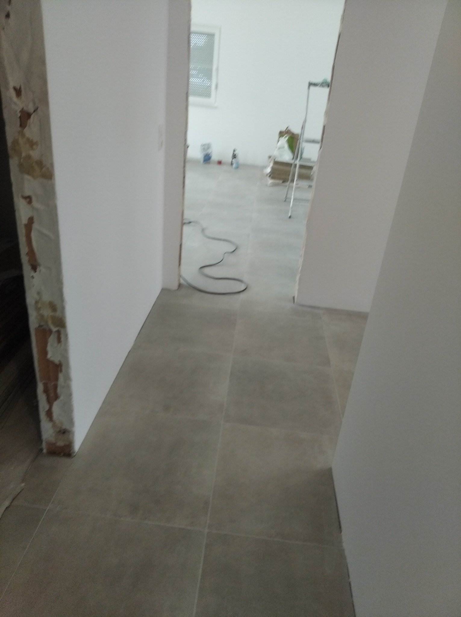 Fragment łazienki w trakcie remontu: widoczne płytki podłogowe w odcieniach szarości, surowe wykończenie ściany z resztkami starej farby, oraz pomieszczenie w głębi z drabiną malarską i materiałami...