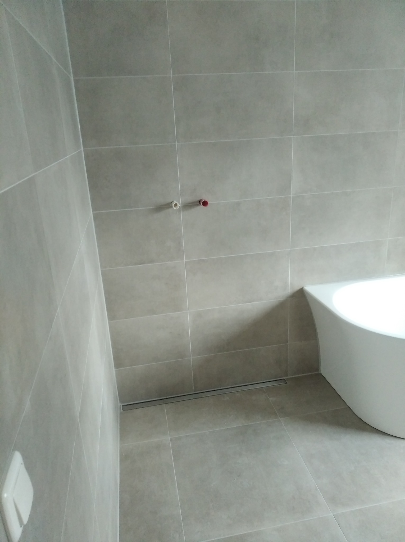 Wykończona łazienka z szarymi, kwadratowymi płytkami, widoczny odpływ liniowy w podłodze i fragment białej wanny, a także wystające rurki z zaworami.