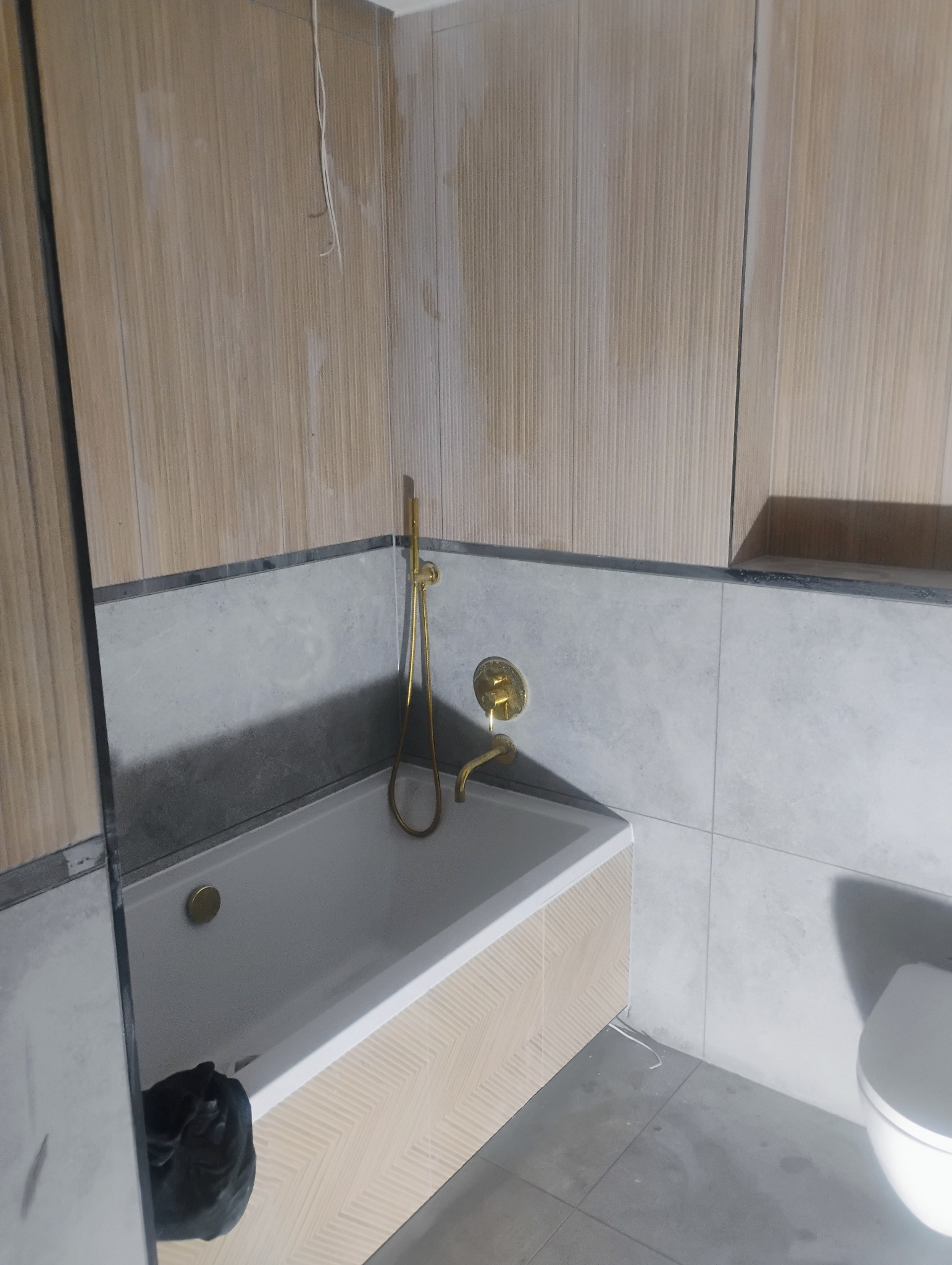 Łazienka w trakcie wykańczania: widoczna wanna z obudową w jasny wzór jodełki, złota bateria, szare płytki na ścianach, częściowo położone płytki imitujące drewno na górze oraz toaleta. Na podłodze...