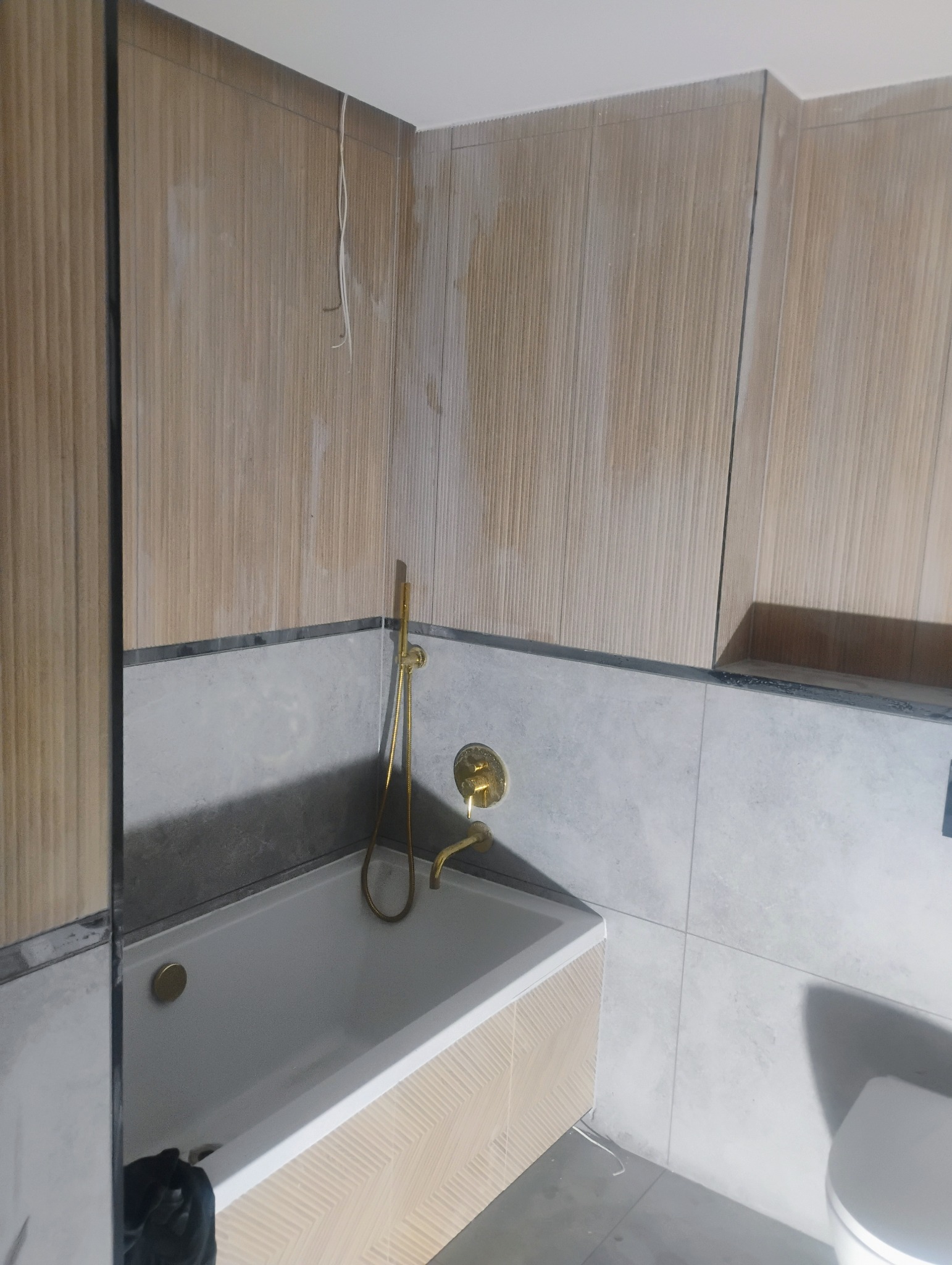 Łazienka w trakcie remontu: obudowana wanna z złotą armaturą na tle częściowo wykończonych ścian z drewnopodobnych i szarych płytek.