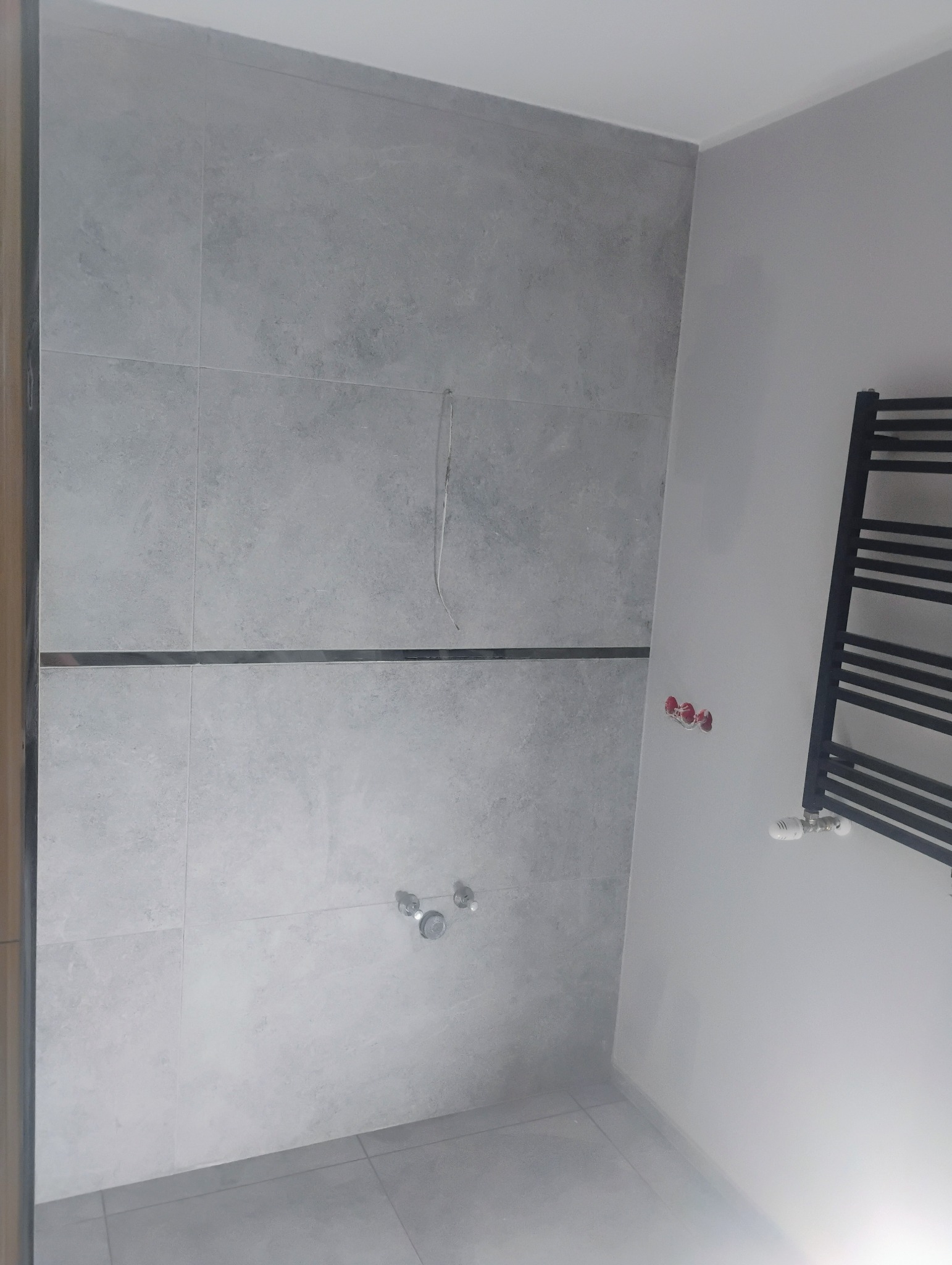 Ściana łazienki wyłożona szarymi płytkami z dekoracyjnym pasem, widoczne przyłącza wodne i grzejnik drabinkowy, instalacja elektryczna w ścianie.