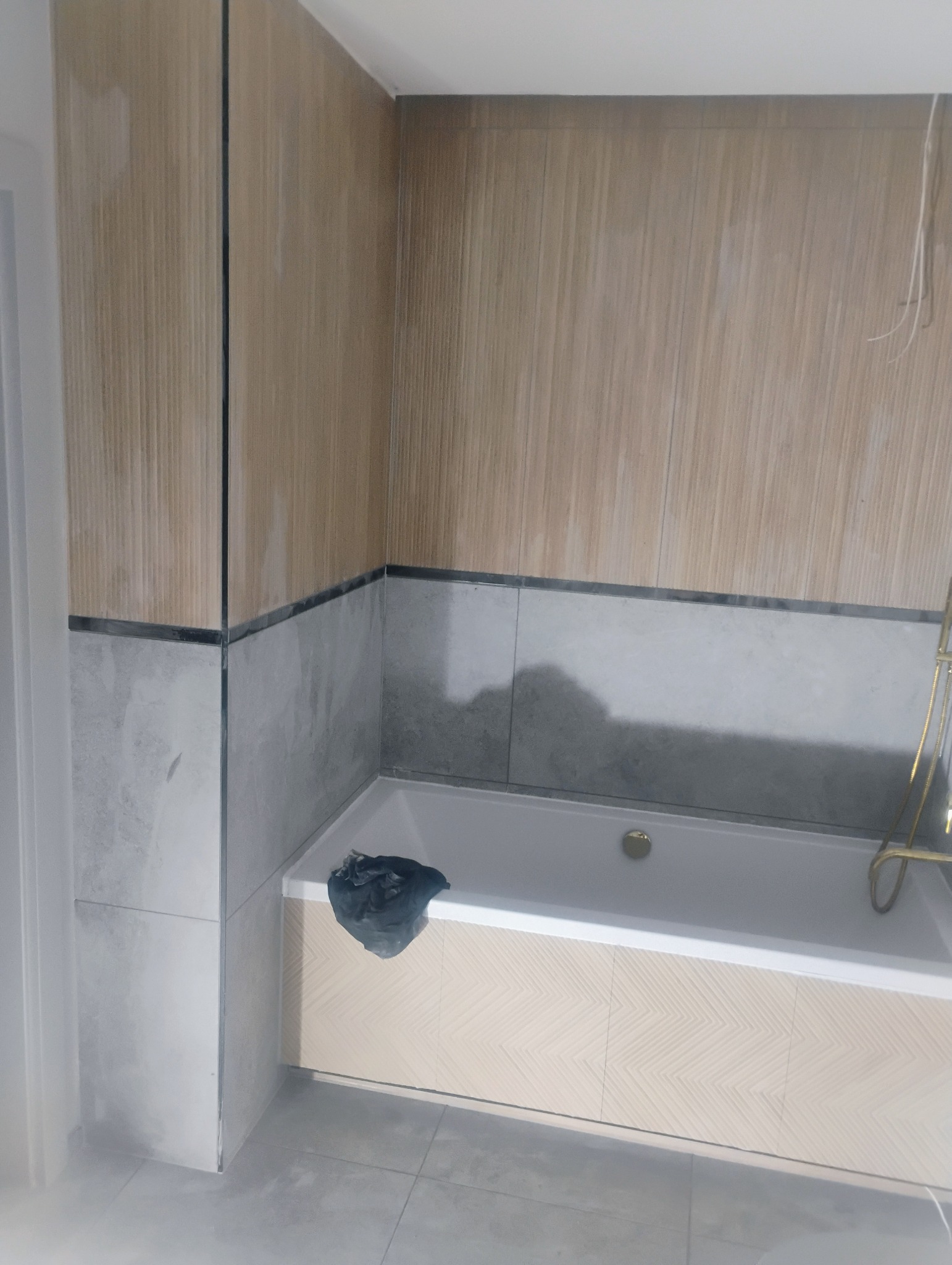 Fragment łazienki w trakcie remontu: położone płytki imitujące drewno na ścianach, szare płytki na dole ścian i podłodze, obudowa wanny z wzorem jodełki, czarny worek na śmieci na wannie.