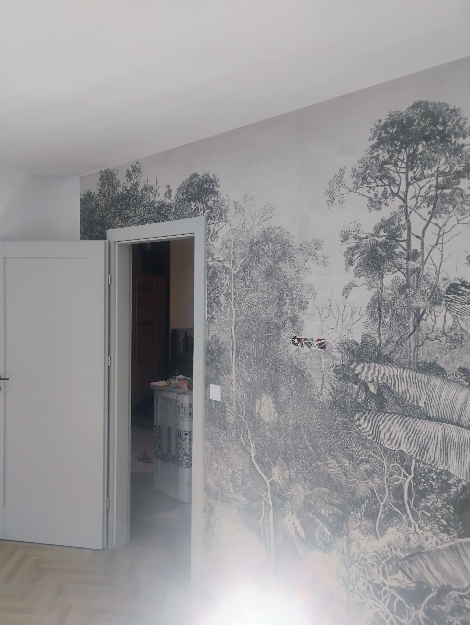 Ściana z fototapetą przedstawiającą grafikę leśną w stylu retro, widoczne niedokończone gniazdko elektryczne z wystającymi kablami, białe drzwi i podłoga z parkietem.