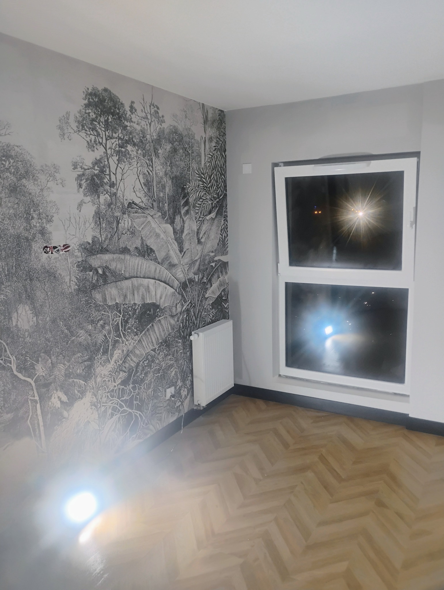 Pokój po remoncie z tapetą w motywy dżungli, otwartym oknem i parkietem ułożonym w jodełkę. Widoczne gniazdka elektryczne bez osprzętu oraz grzejnik.