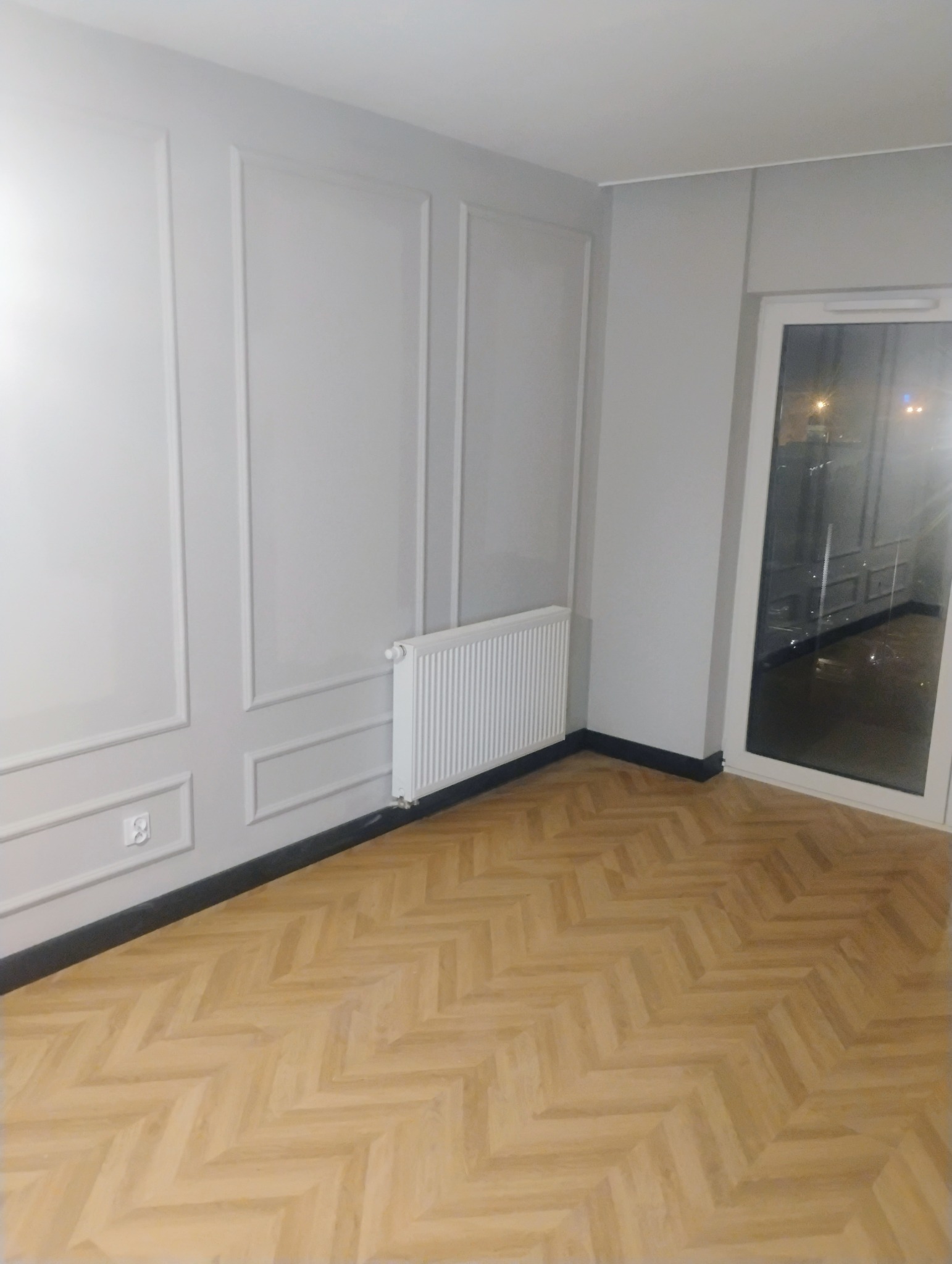 Pusty pokój po remoncie z nową podłogą w jodełkę, pomalowanymi na szaro ścianami z dekoracyjnymi listwami i białym grzejnikiem.