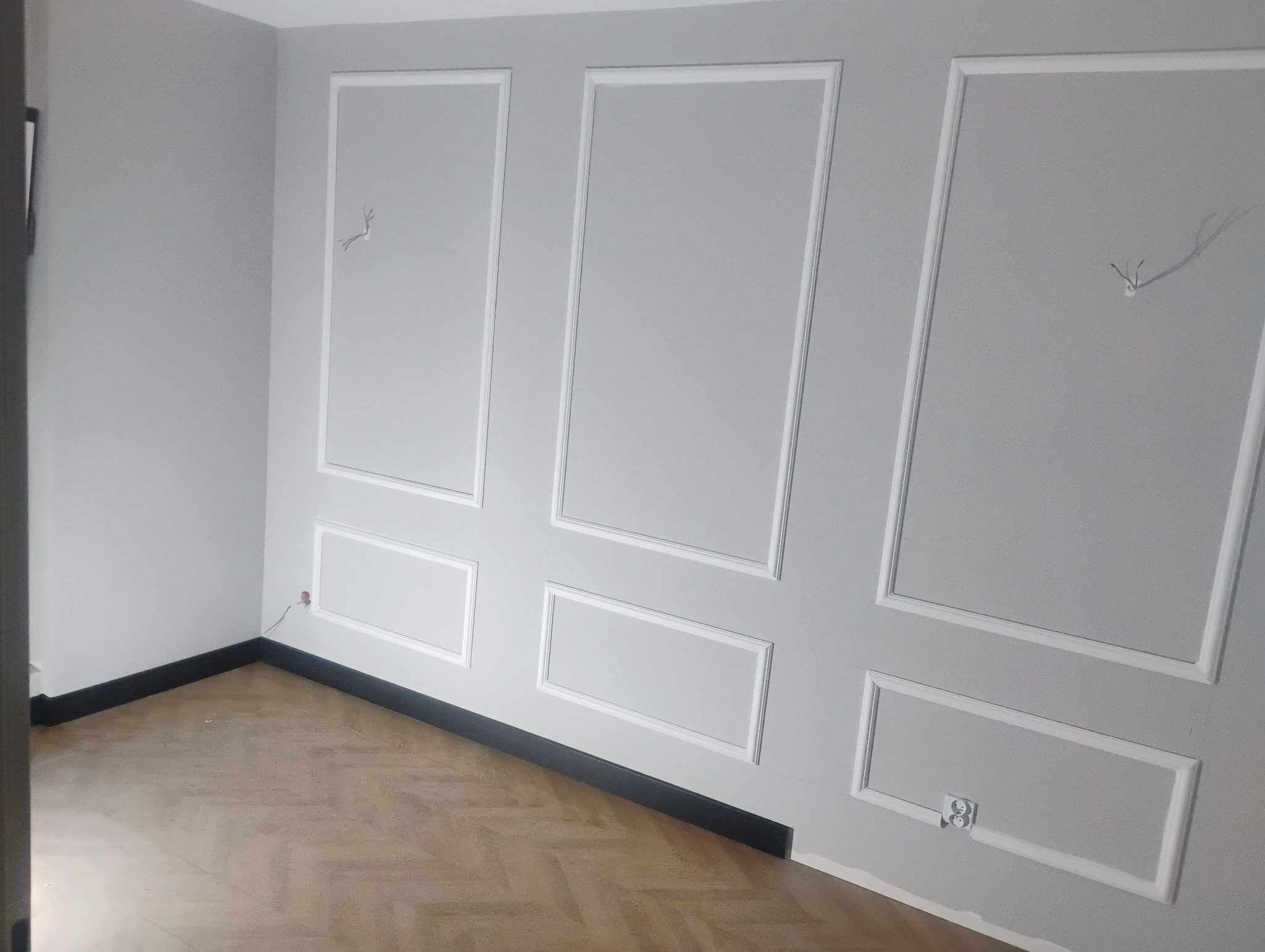 Szare wnętrze pokoju z panelami ściennymi w białych obramowaniach, parkiet w jodełkę, czarna listwa przypodłogowa i wystające kable ze ściany.