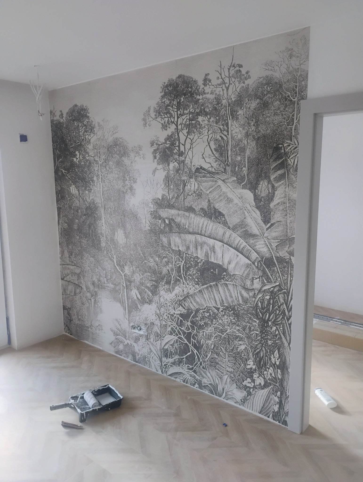 Ściana z fototapetą przedstawiającą dżunglę w stylu retro, na podłodze wałek do malowania i butelka kleju, widoczne niedokończone prace wykończeniowe.