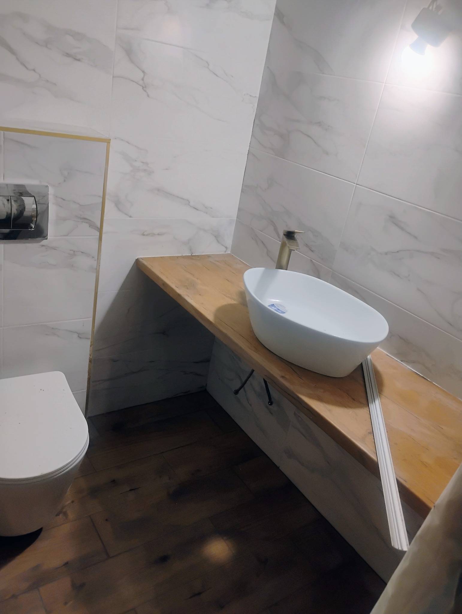 Nowoczesna łazienka z marmurowymi płytkami, drewnianym blatem pod umywalką nablatową w kształcie elipsy i ciemną drewnianą podłogą. Widoczna toaleta i bateria umywalkowa w kolorze szczotkowanego...