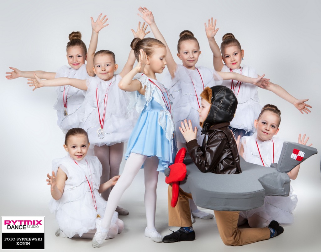 Grupa dzieci w kostiumach tanecznych i z medalami, jedno dziecko przebrane za pilota w tekturowym samolocie, na jasnym tle.