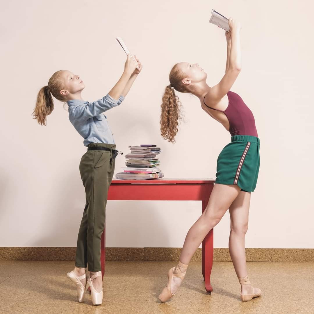 Dwie baletnice w strojach do ćwiczeń, jedna w jeansowej koszuli i spodniach, druga w bordowym body i zielonych szortach, ćwiczą postawę z książkami nad głowami, stojąc obok czerwonego stolika...