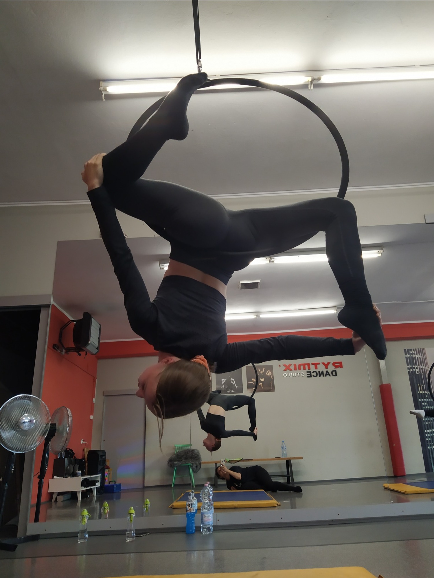 Młoda kobieta w czarnym stroju wykonuje figurę akrobatyczną na aerial hoop w studiu tańca, widoczne odbicie w lustrze i logo studia Rytmix Dance Studio.