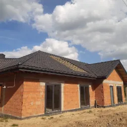 Niedokończony dom jednorodzinny z czerwonej cegły z zamontowanymi oknami i częściowo położonym ciemnobrązowym dachem na tle błękitnego nieba z białymi chmurami.