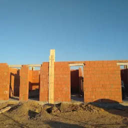 Wznoszenie ścian budynku z czerwonej cegły ceramicznej, widoczne otwory okienne i drzwiowe, drewniane stemple podtrzymujące nadproża, nasyp piasku przed budynkiem w słoneczny dzień.