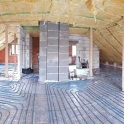 Instalacja ogrzewania podłogowego w trakcie montażu na poddaszu, widoczne niebieskie rurki na siatce, ocieplenie wełną mineralną na skośnym dachu, surowy stan wykończenia ścian.