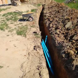 Wykop w ziemi z ułożonymi niebieskimi rurami, widoczna taczka i budynek w tle, słoneczny dzień.