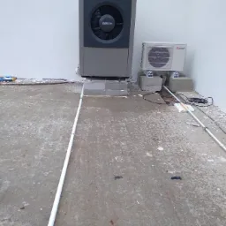 Dwa urządzenia grzewcze, Buderus i Mitsubishi, zamontowane na betonowych bloczkach na surowym betonowym podłożu przy białej ścianie. Widoczne rury instalacyjne prowadzące do urządzeń.