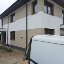 Dwukondygnacyjny dom z ocieploną elewacją w odcieniach beżu i bieli, z brązowymi ramami okiennymi, częściowo zasłonięty białym samochodem dostawczym z otwartymi drzwiami.