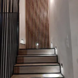 Schody z drewnopodobnymi panelami i czarnymi obramowaniami stopni, oświetlone małymi lampkami LED w ścianie i pod stopniami, obok dekoracyjna ścianka z pionowych, drewnianych listew.