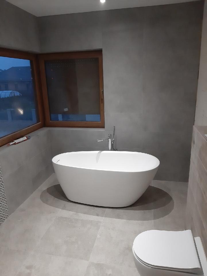 Nowoczesna łazienka z wolnostojącą białą wanną, szarą glazurą ścienną i podłogową, drewnianą ramą okienną i podwieszaną toaletą