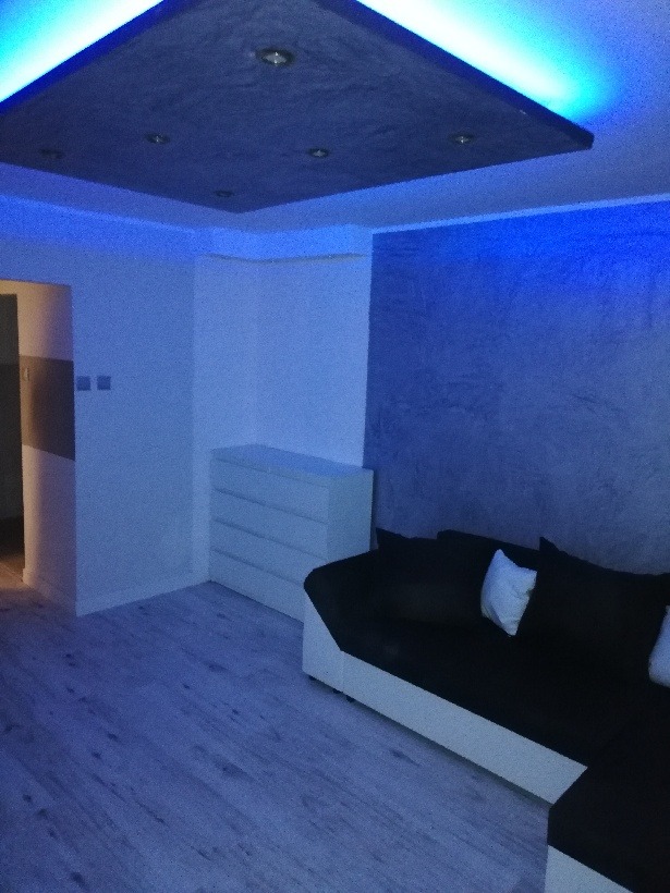 Pokój z szarą, teksturowaną ścianą oświetloną niebieskim światłem LED, szary sufit z wbudowanymi punktami świetlnymi, biała komoda i czarno-biała sofa narożna